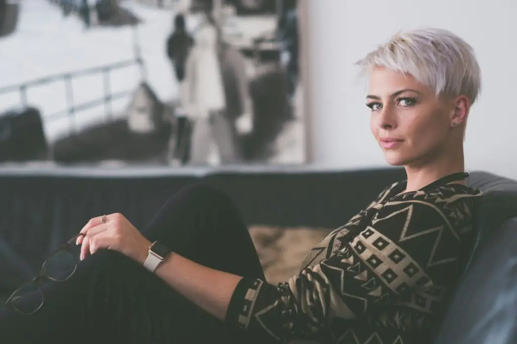 Woman with short blonde hair sitting calmly on a couch, holding glasses, looking thoughtfully toward the camera.