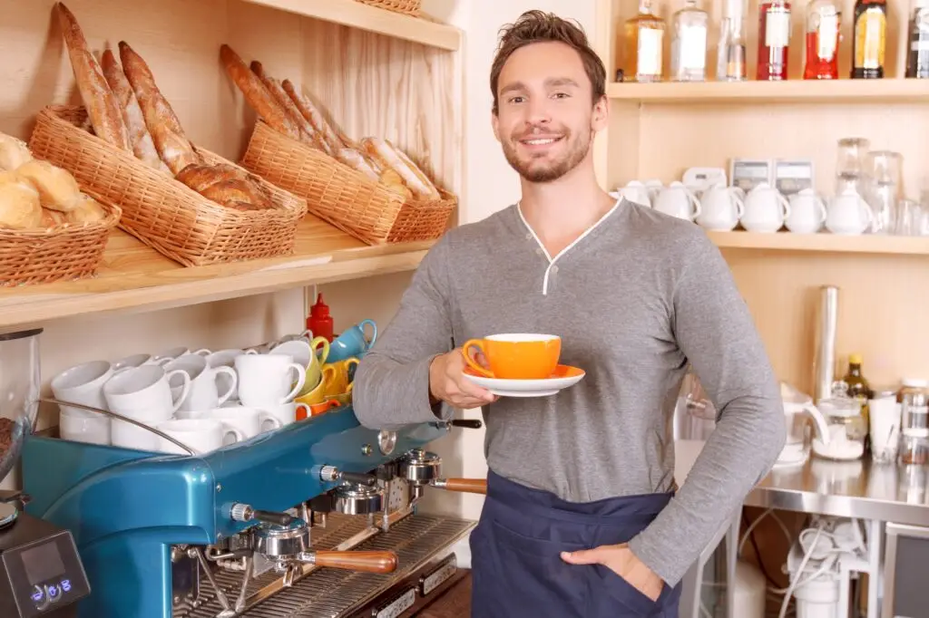 Smiling barista holding an orange coffee cup in a cafe with bread baskets and an espresso machine.