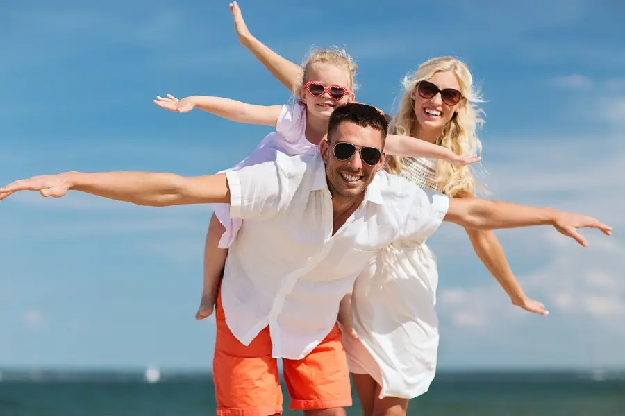 Happy family with arms outstretched smiling together on a sunny beach under a blue sky.