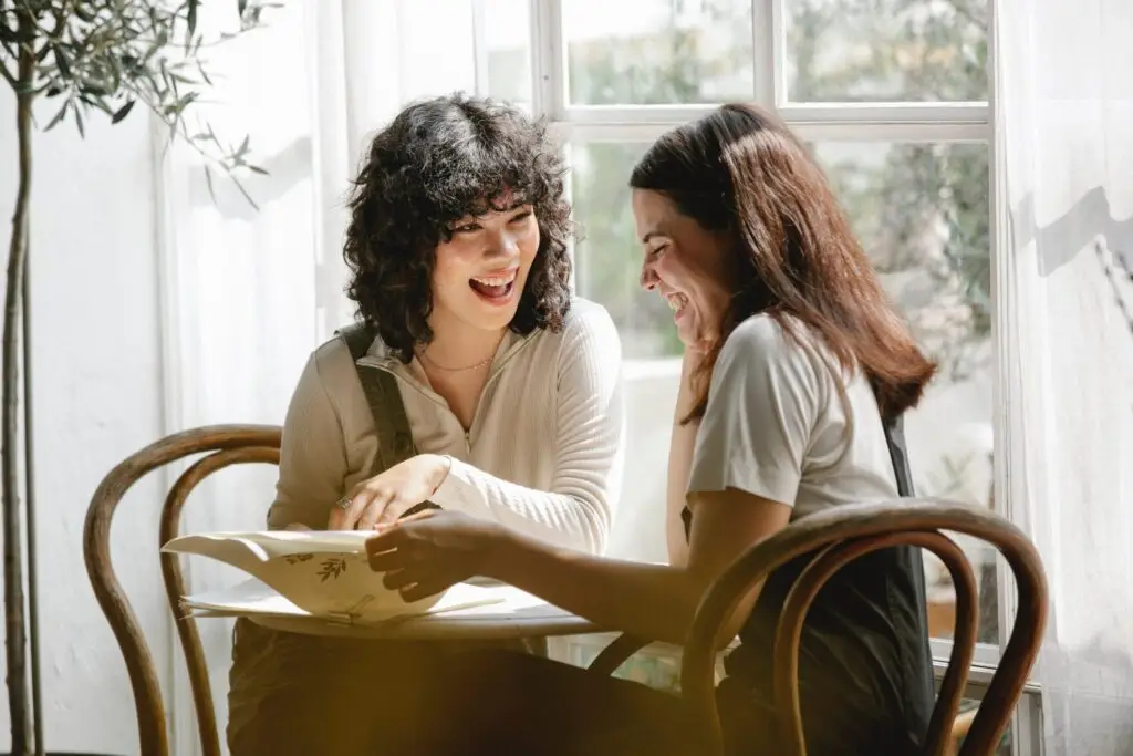 Two women laughing together at a sunlit table while sharing a journal, supporting each other in recovery.